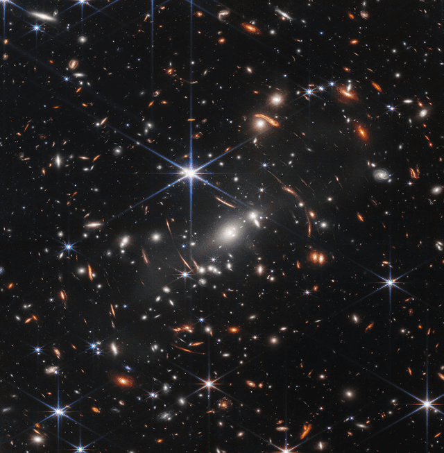 A black field crowded with thousands of galaxies and stars; orange arcs curve around the center where gravity lenses distant light.
